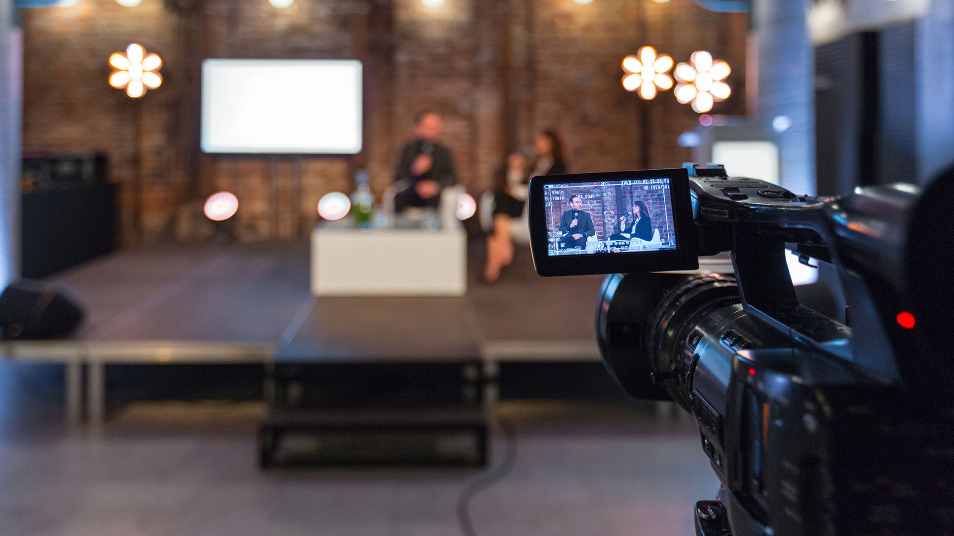 Businesswoman and businessman giving presentation during online seminar, sitting on armchairs on the stage, holding microphones. Focus on video camera on the foreground.