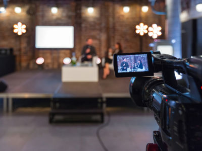 Businesswoman and businessman giving presentation during online seminar, sitting on armchairs on the stage, holding microphones. Focus on video camera on the foreground.